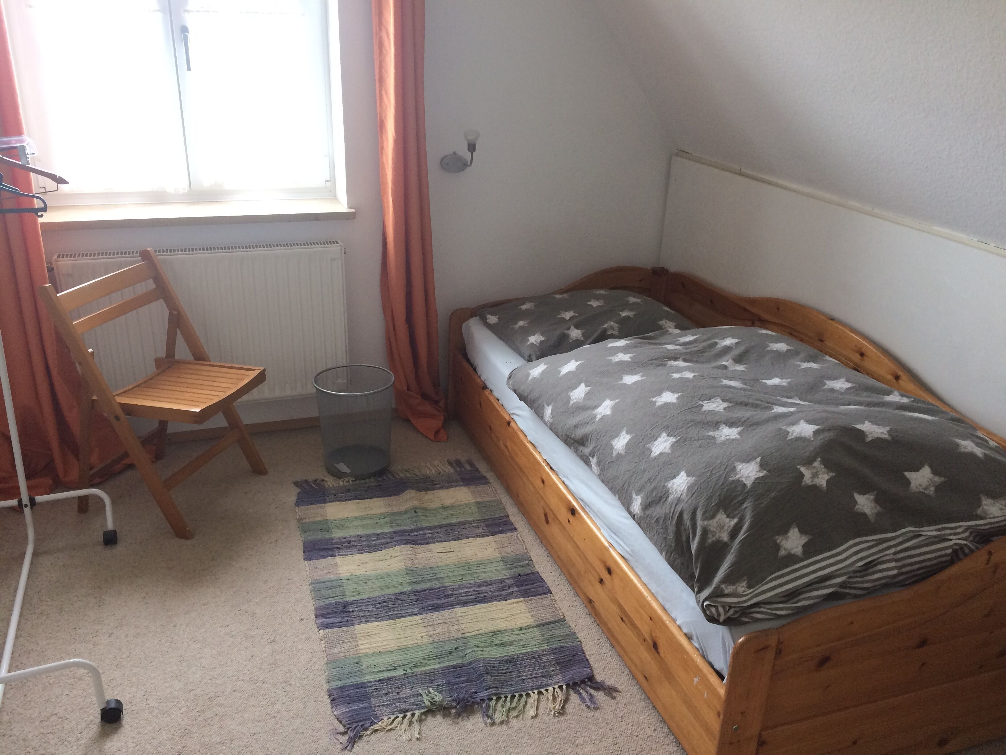 Une petite chambre avec un lit simple, une tête de lit en bois, une couverture à motifs étoilés, un tapis à rayures, une chaise et un radiateur.