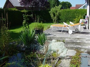 Un jardín con un patio de piedra, sillas de lounge y diversas plantas.