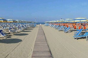 Ein Strand mit Liegestühlen, Sonnenschirmen, einer Holzpromenade und blauem Himmel.