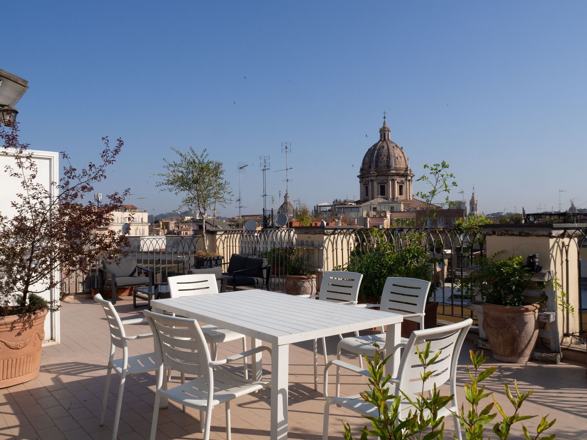 Una terraza en la azotea con una mesa y sillas blancas, plantas en macetas y vistas a un edificio histórico con cúpula.