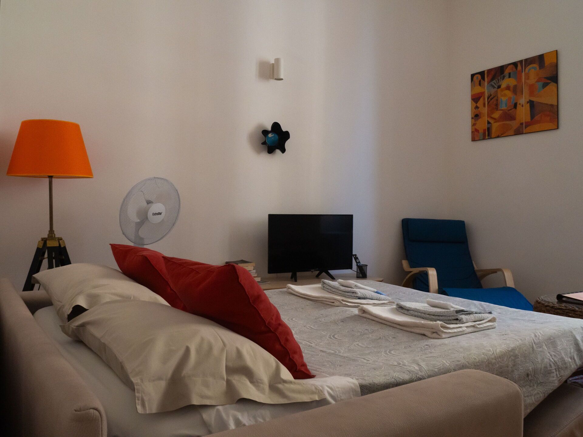 A bedroom with a bed, a desk, a chair, a lamp, a fan, and a painting on the wall.