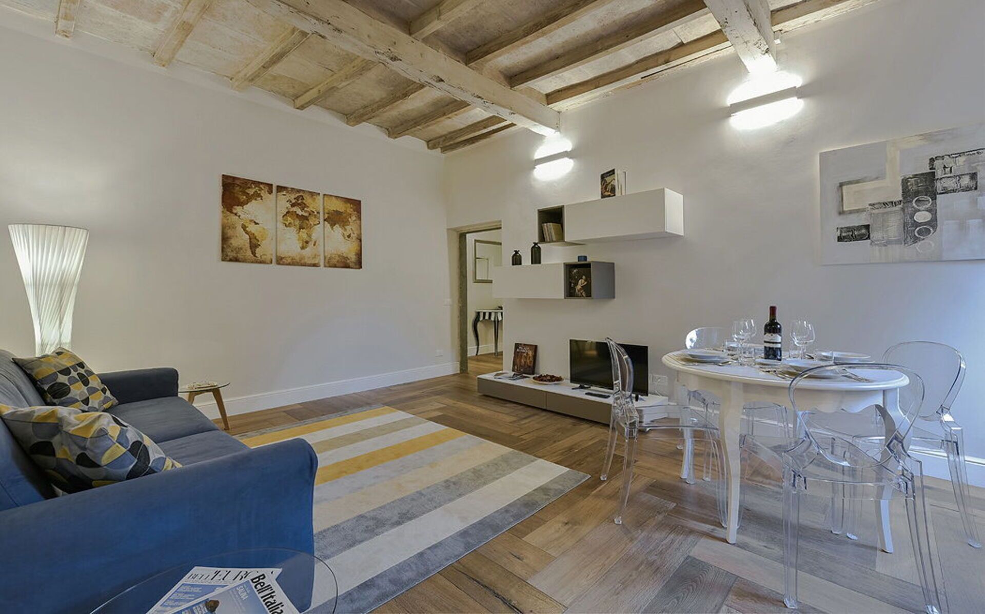 Ein Wohnzimmer mit einem blauen Sofa, einem Streifen Teppich, einem Esstisch mit transparenten Stühlen und einer Weltkarte an der Wand.