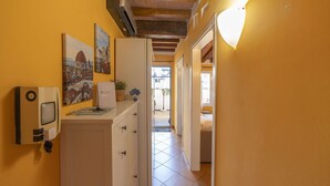 Een smalle gang met een wit kastje, een apparaat aan de muur en een deur naar een kamer met een bed.
