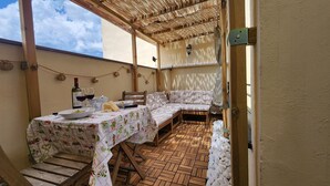 Een overdekt terras met een houten eettafel, een zithoek met kussens en uitzicht op de lucht.