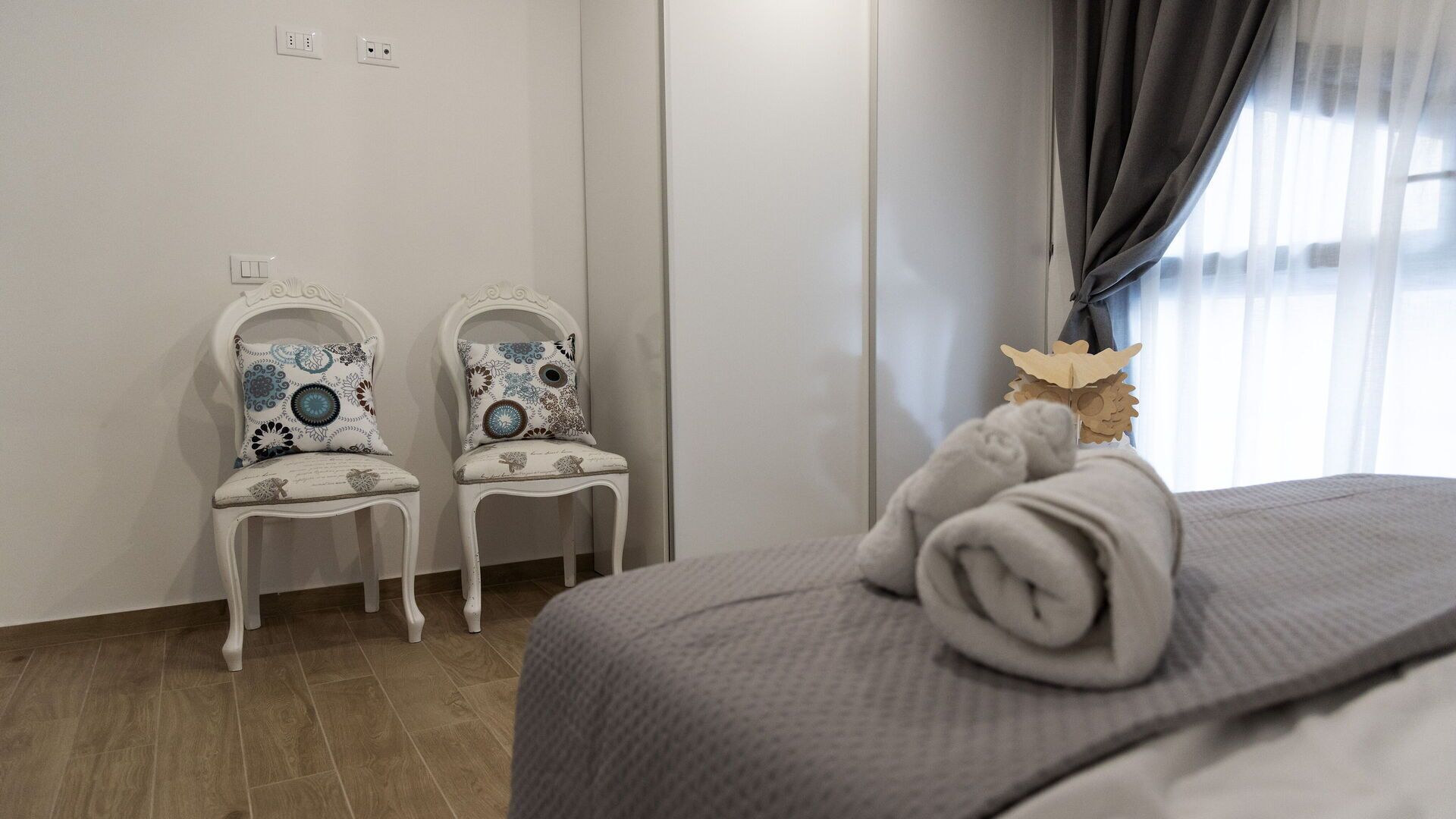 A hotel room with a bed, two chairs with patterned cushions, a window with curtains, and a wall socket.