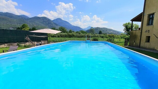 A clear blue swimming pool with a mountain backdrop, surrounded by a fenced area and a building with a sloped roof.