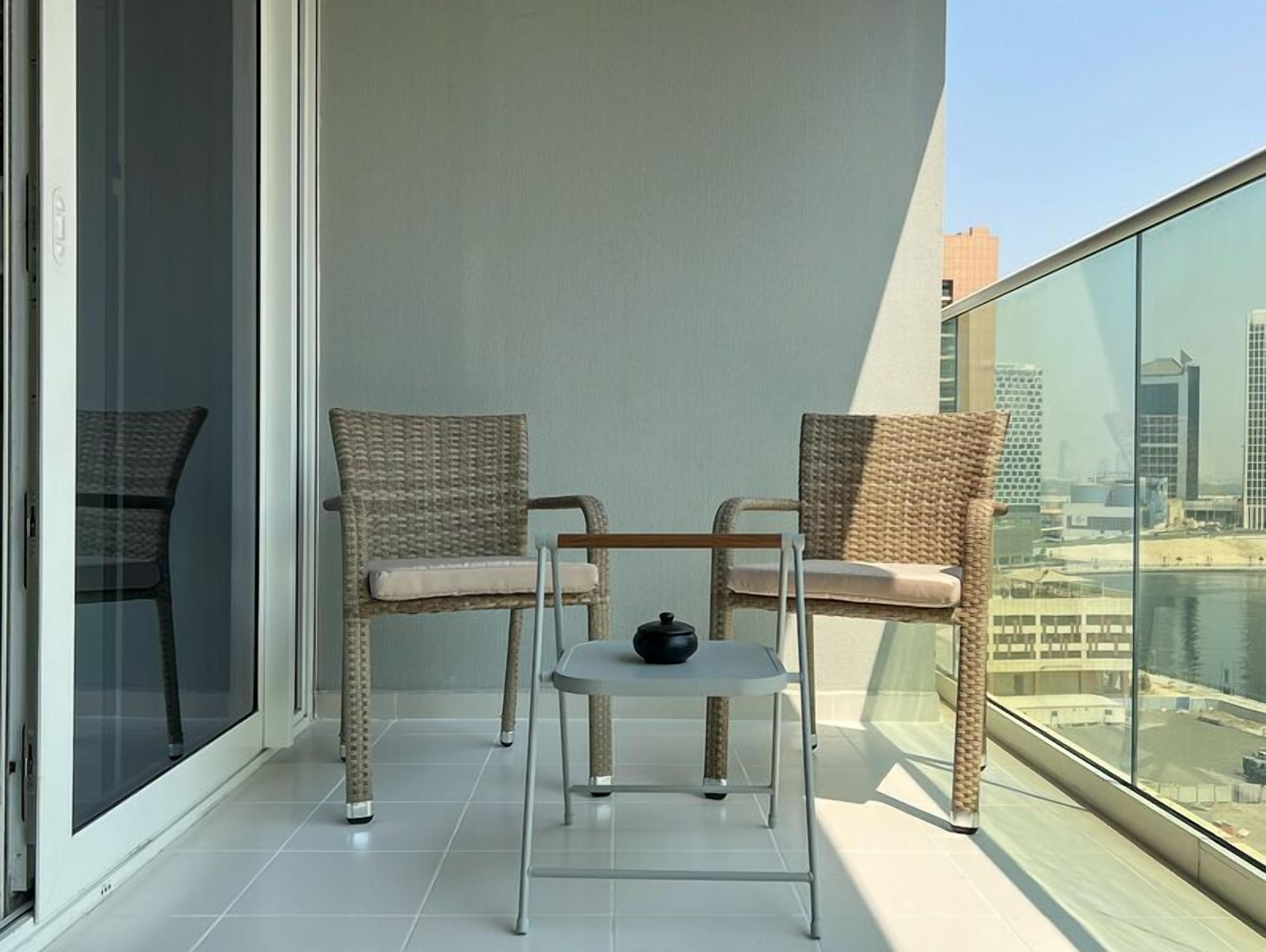 A balcony with two wicker chairs and a small table, overlooking a cityscape.