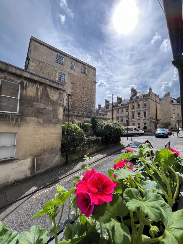 A street view with a stone building, a parked car, and a flower bed with pink flowers. - Cosy Georgian cottage, very central, newly renovated, with a courtyard, sleeps 3 (Bath)