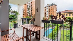 A balcony with a wooden table and chairs, potted plants, and a view of a swimming pool and surrounding buildings. - City Oasis · Terrace and swimming pool (Milano)