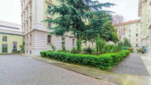 Une cour avec un grand arbre, une haie et des bâtiments à l’architecture classique.