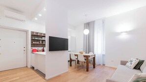 Un salon moderne avec un plancher en bois, un coin repas avec une table et des chaises, un canapé avec des coussins, un téléviseur à écran plat fixé au mur et un coin cuisine avec des étagères et un réfrigérateur.