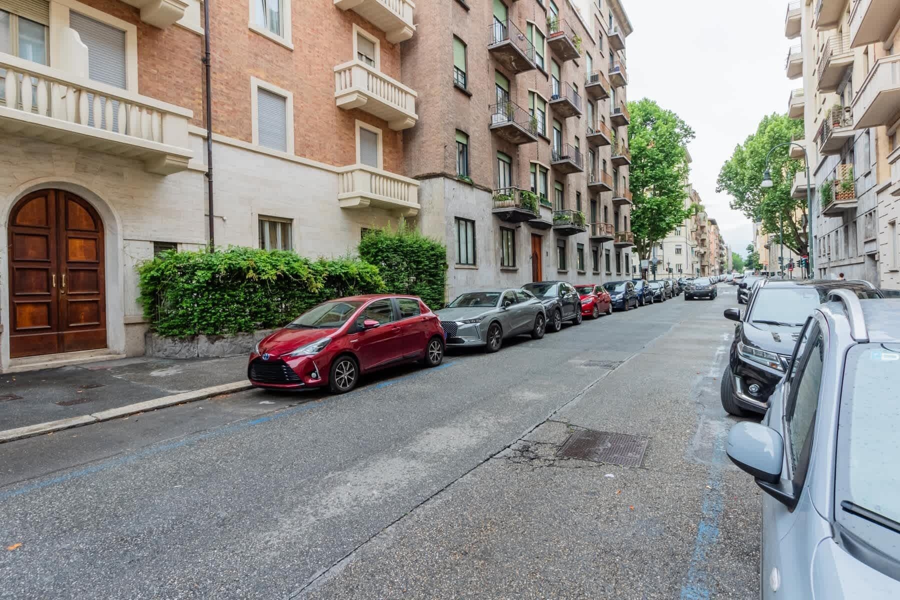 Una fila de coches estacionados a lo largo de una calle residencial con edificios de varios pisos, un coche rojo al frente y una puerta de madera grande a la izquierda.