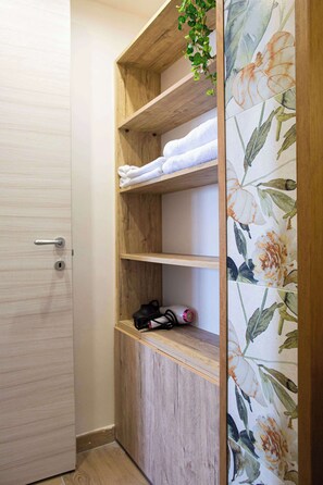 A corner of a room with a wooden shelf holding towels and a pair of shoes, a door with a handle, and a wall with floral wallpaper.