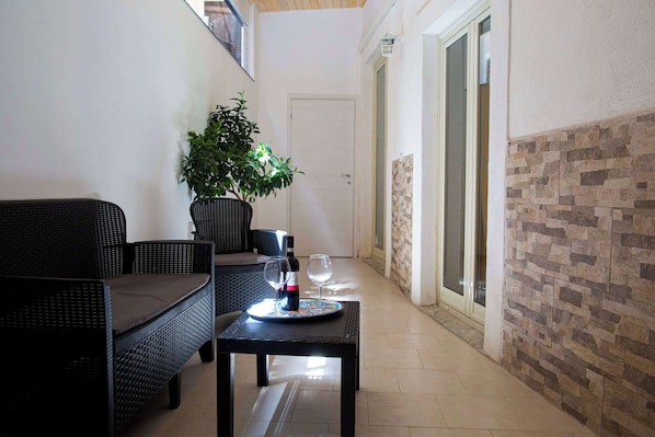 A corridor with a wicker sofa, a small table with a bottle and glasses, a potted plant, and a door leading to another room.