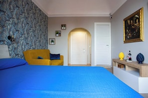 A bedroom with a bed, a yellow armchair, a desk, a door, and a wall with a patterned wallpaper and a framed painting. - Charming Studio Verga, by the old town (Catania)