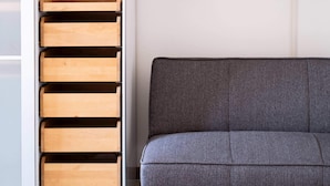 Una habitación con un sofá gris, un televisor montado en la pared y un estante blanco con cajones de madera.