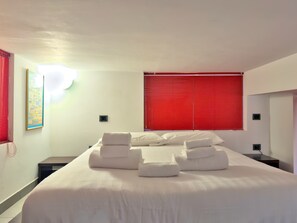 Una cama bien hecha con ropa de cama y toallas blancas, una persiana roja y una mesita de noche con lámpara.