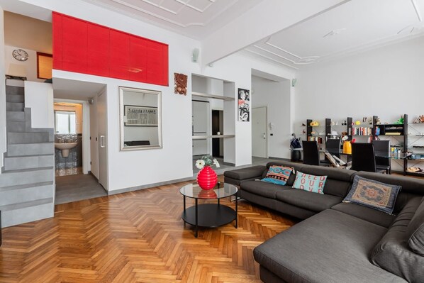 Un salón moderno con suelo de madera en espiga, un sofá de cuero negro, una mesa de centro de cristal y un jarrón rojo.