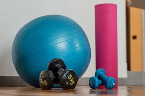 Una pelota de ejercicios azul, mancuernas negras y azuladas, y un objeto cilíndrico rosa sobre un piso de madera.