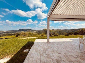 A covered outdoor area with a brick floor, a white chair, and a view of a grassy landscape with hills in the distance.