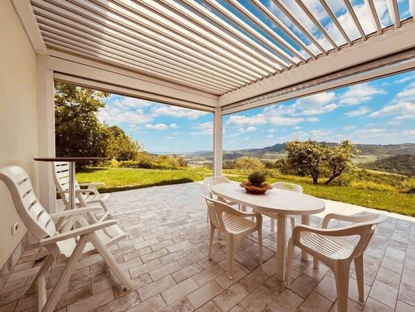 An outdoor patio with a table and chairs, a pergola, and a view of a green landscape.
