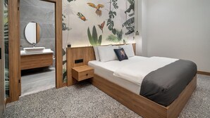 Une chambre avec un cadre de lit en bois, un matelas blanc, une couverture grise, une table de chevet, une commode en bois, un miroir et un mur orné d’une fresque tropicale.