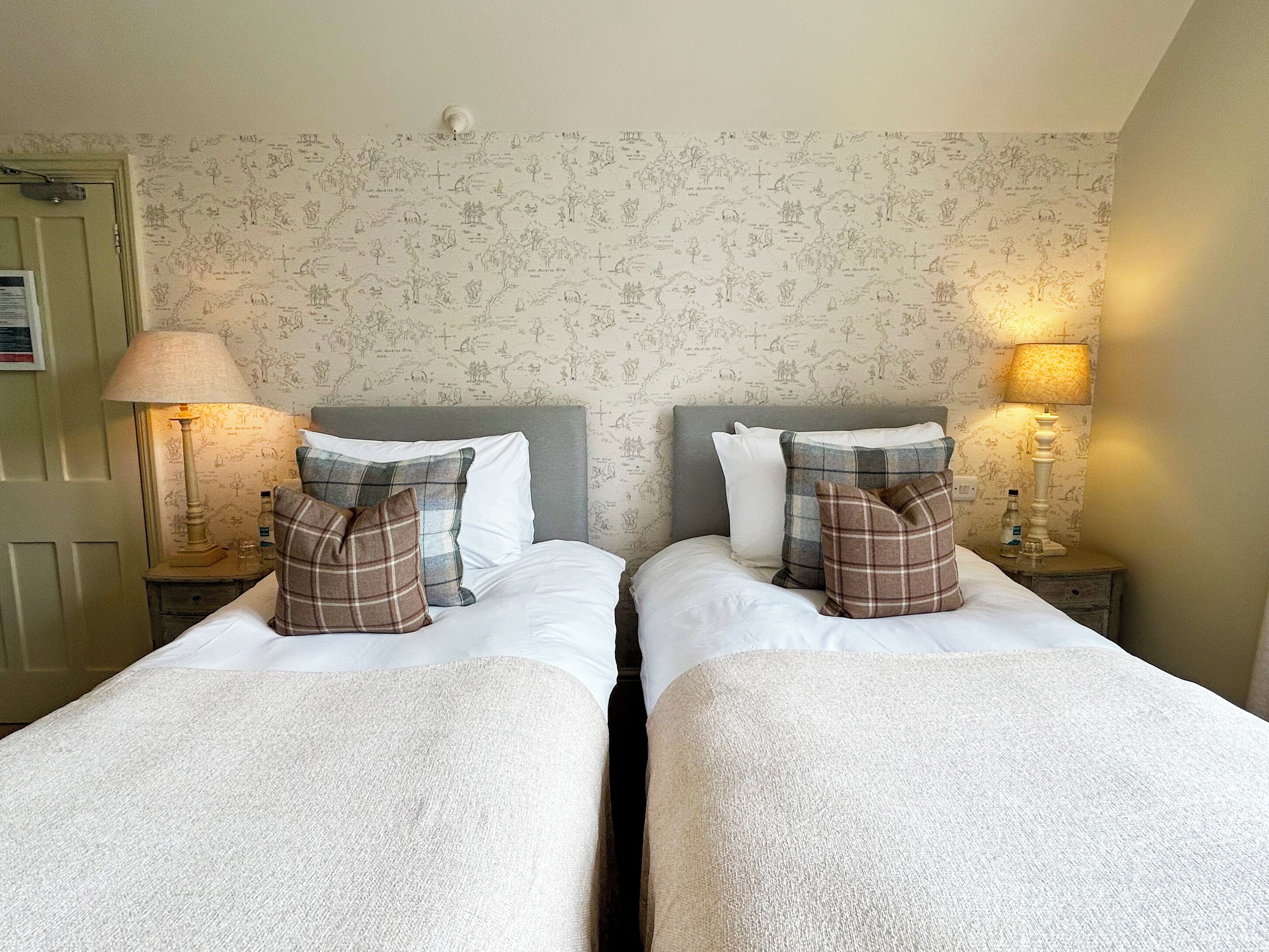 A double room with two beds, each with a grey headboard, white bedding, and plaid pillows. There are bedside lamps and a wall with a map-like pattern.