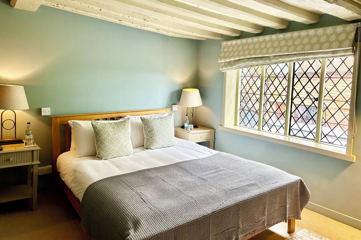 A bedroom with a bed, two bedside tables, a window with a grid pattern, and a wooden ceiling.