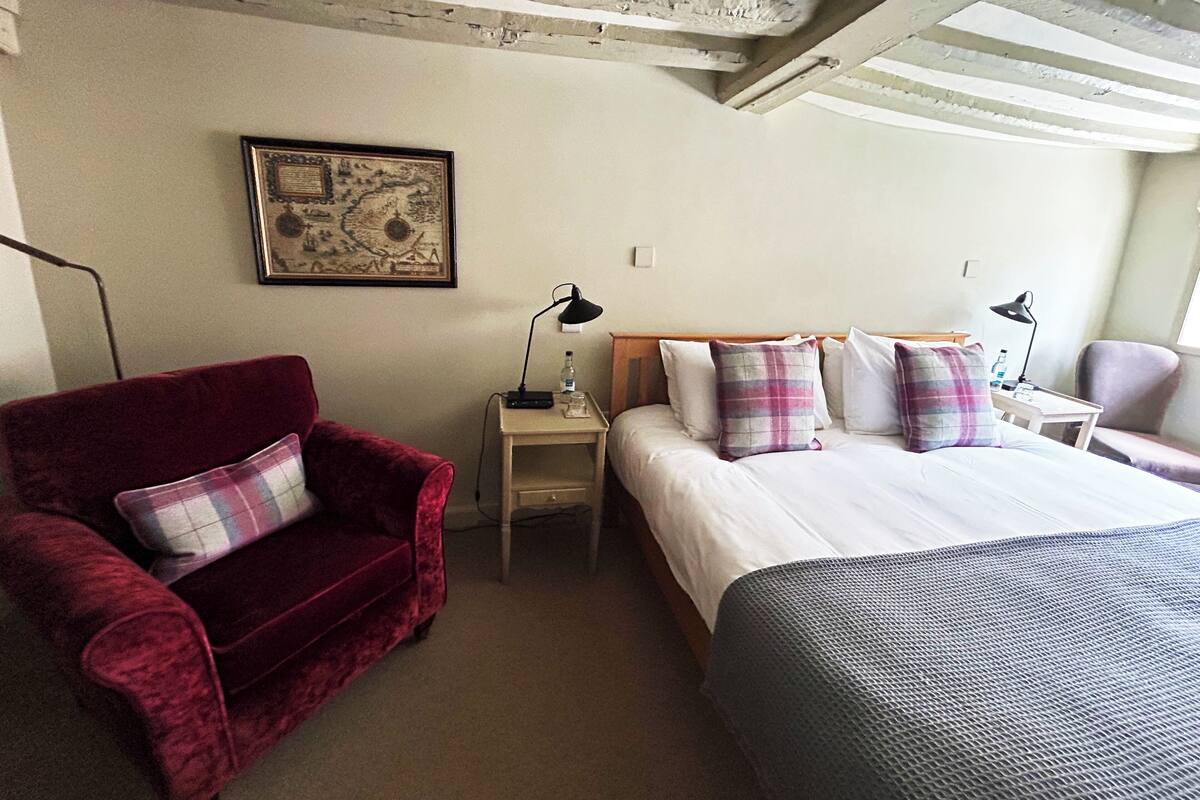 A hotel room with a red velvet armchair, a bed with a grey blanket, a wooden headboard, and a nightstand with a lamp and a bottle.