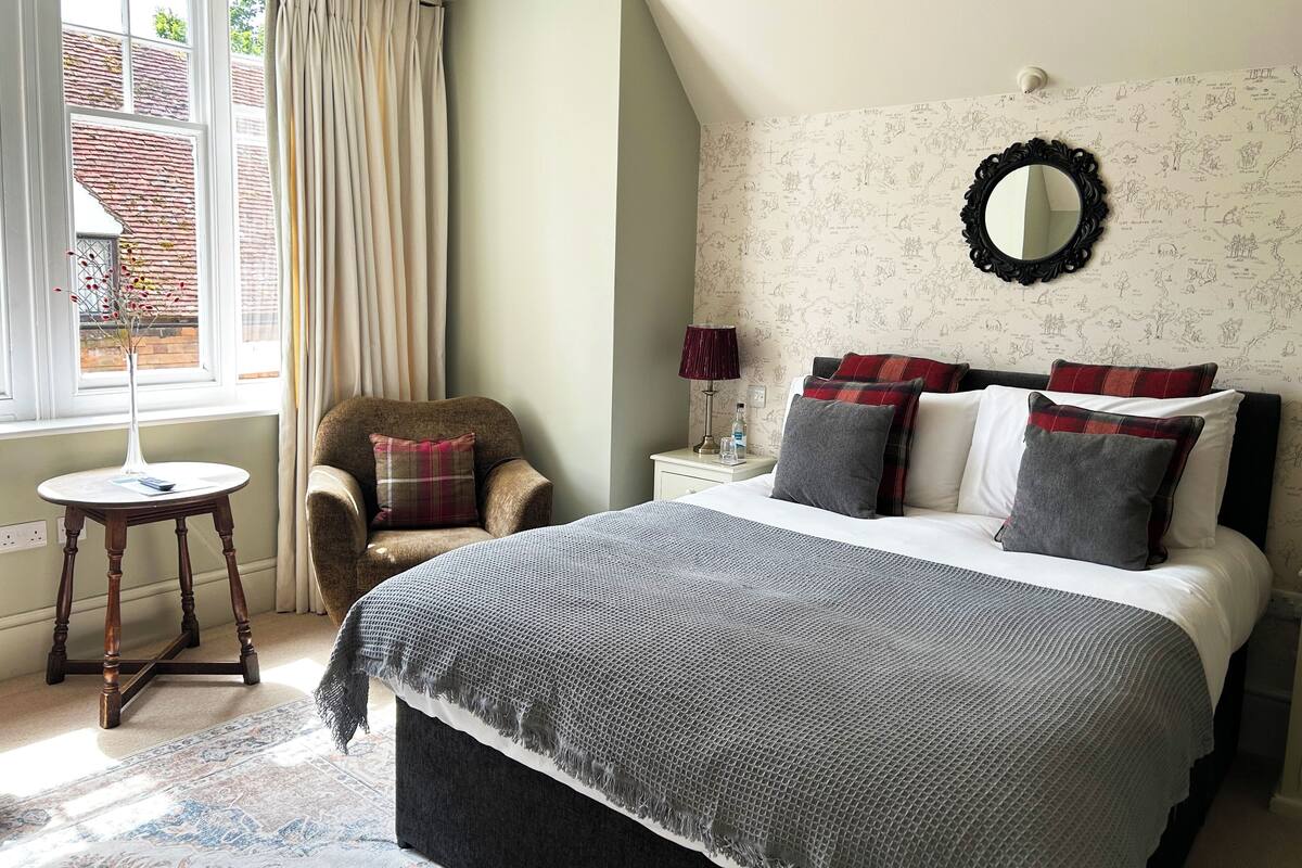 A bedroom with a bed, a chair, a nightstand, a lamp, and a window with curtains.