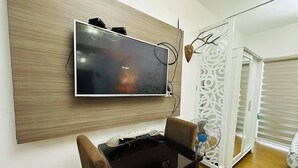 A modern living room with a flat-screen TV mounted on a wooden panel, a small table with a keyboard, and a decorative fan. - Affordable stays in taguig near bgc (Taguig)