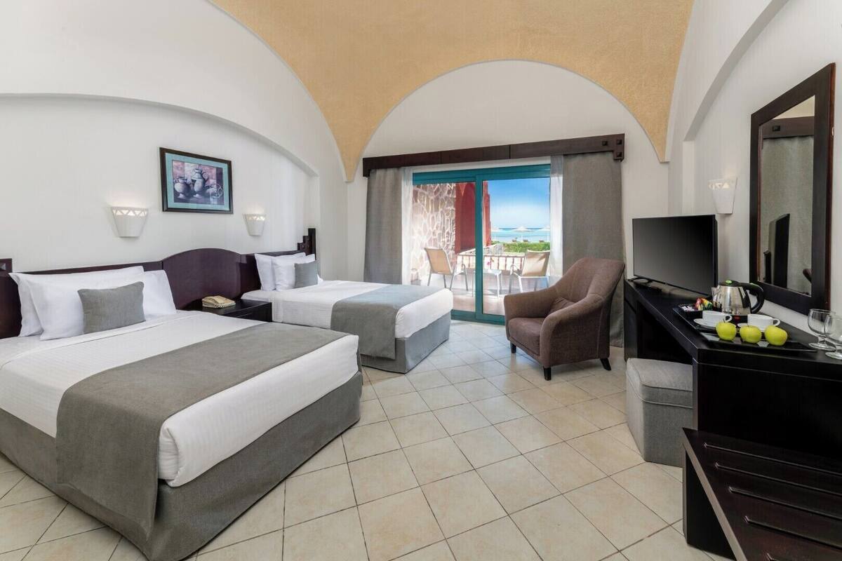 Habitación de hotel con dos camas, televisión, escritorio y bandeja de frutas, y vista al mar.