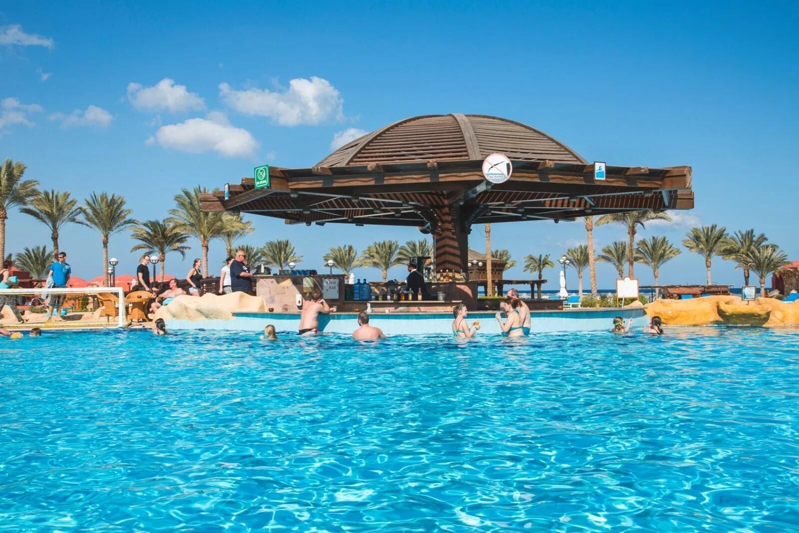 A swimming pool with a wooden pavilion, palm trees, and a clear sky.