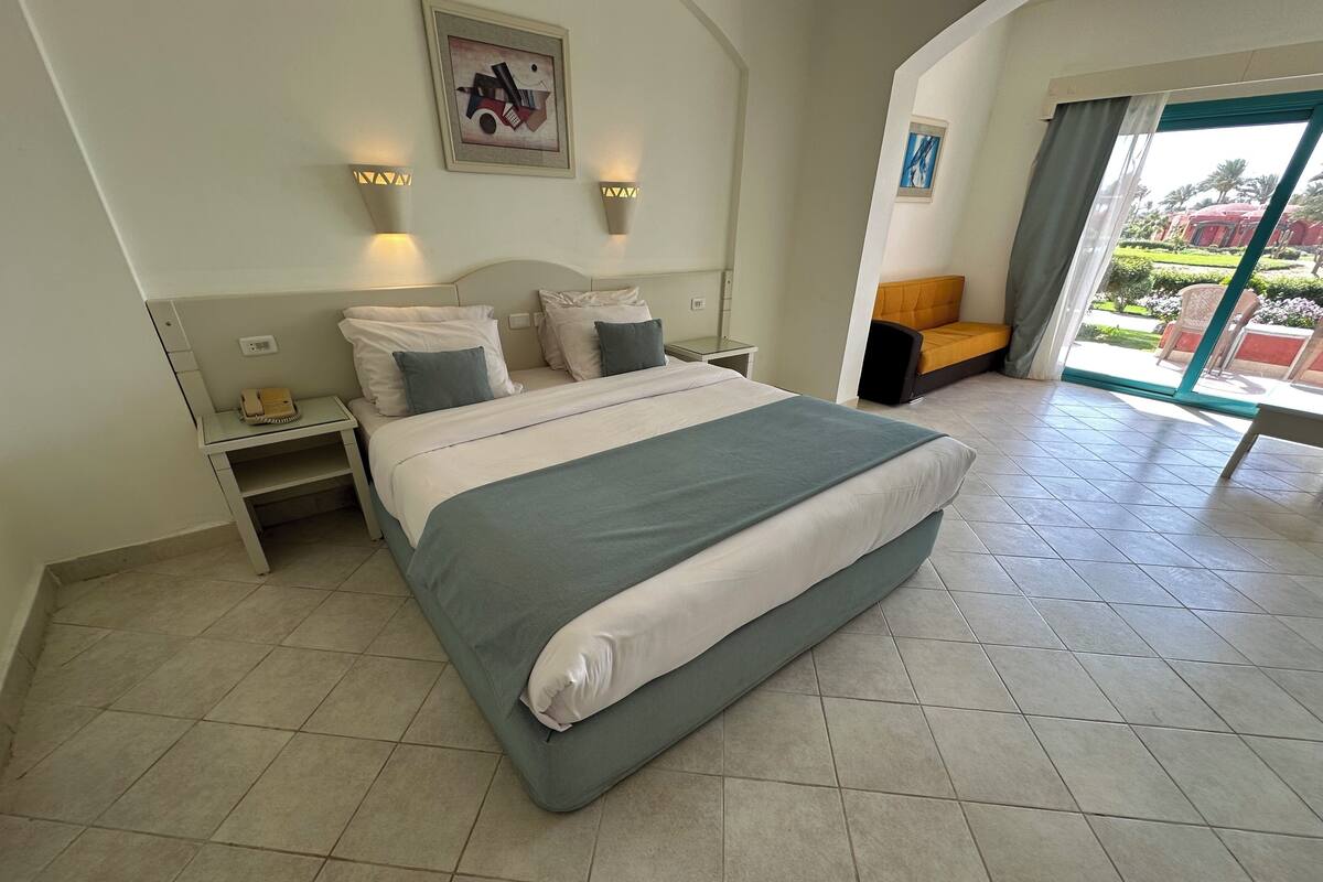 Habitación de hotel con una cama grande, dos mesitas de noche, una silla, un ventanal con vistas y un cuadro en la pared.