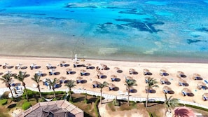 Eine Vogelperspektive eines Strandes mit Reihen von Liegestühlen und Sonnenschirmen, Palmen und einem klaren, blauen Meer.