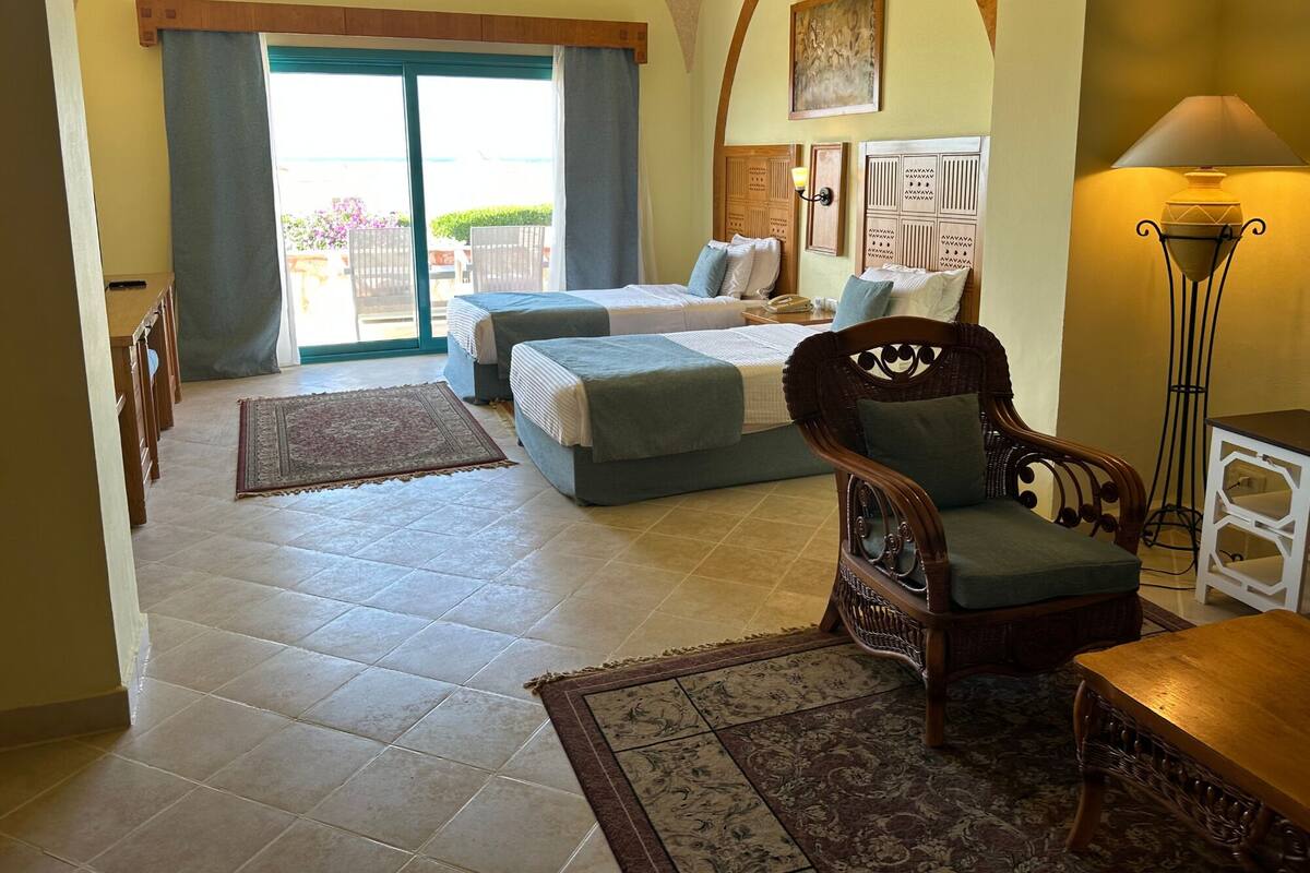 Una habitación con dos camas, una mesa de comedor, una silla, una lámpara y vistas al exterior.