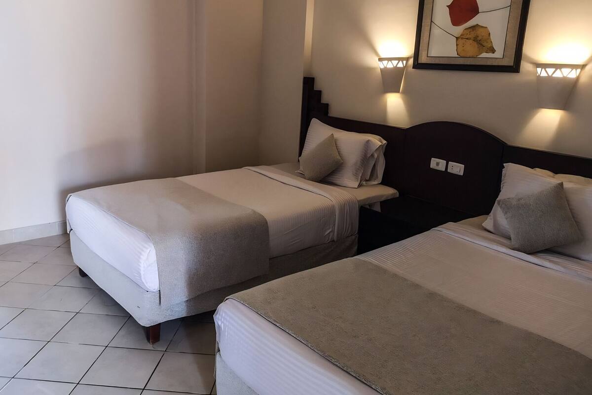 Habitación de hotel con dos camas, cabecero, luces fijadas a la pared y un cuadro enmarcado.
