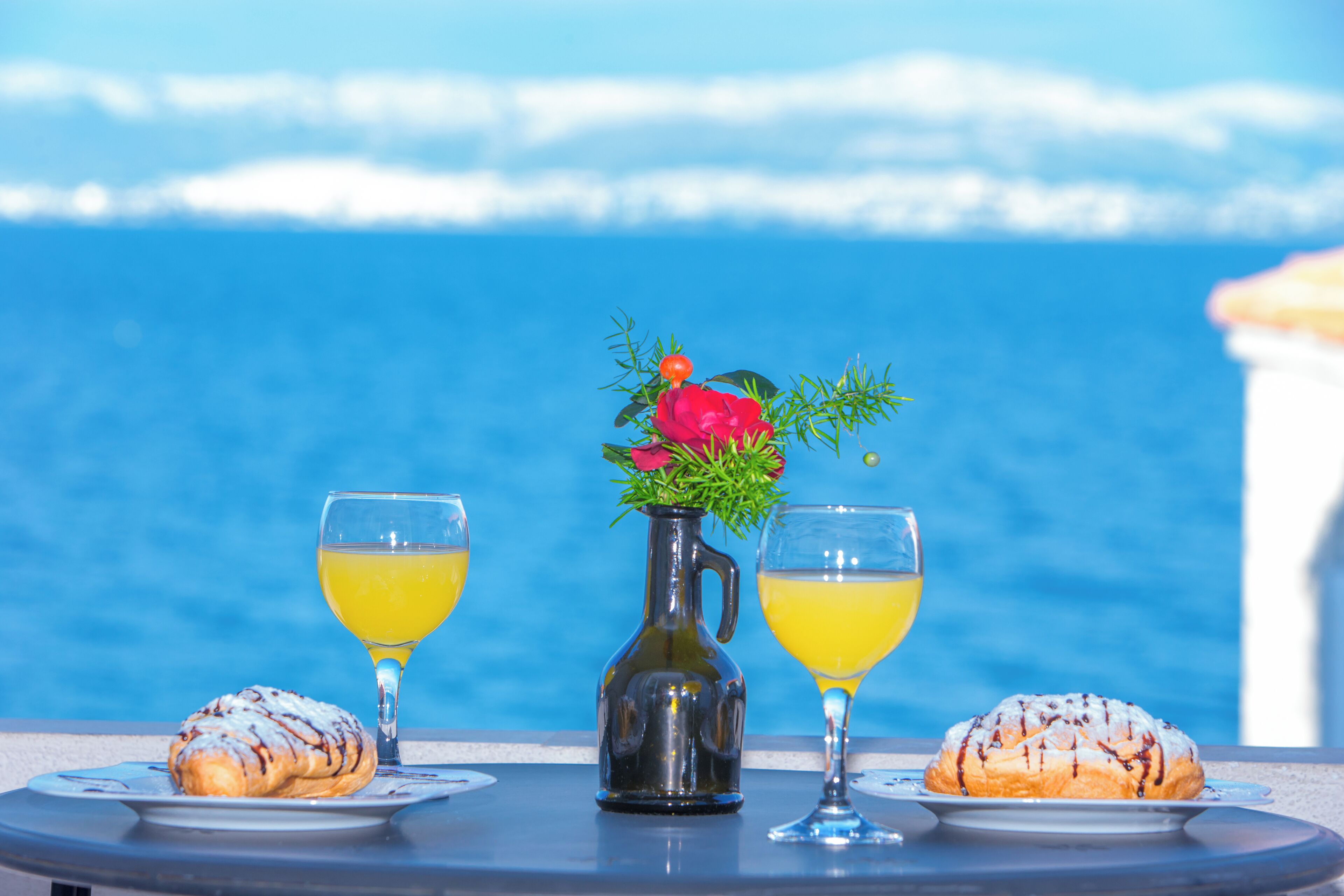 A table with two pastries and two glasses of yellow beverage, a vase with flowers, and a view of the sea.