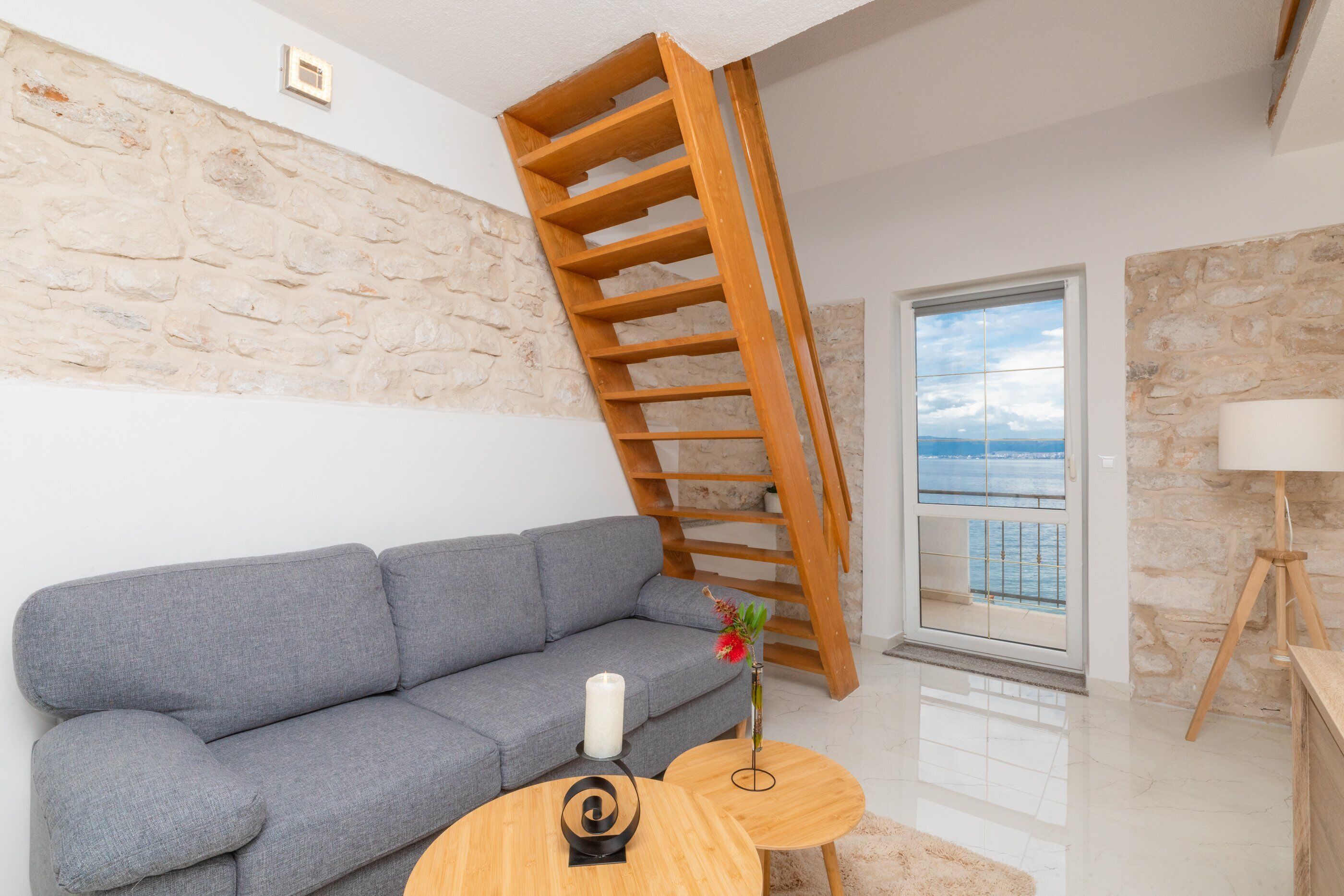 Un salon moderne avec un canapé gris, une échelle en bois, une petite table avec une bougie et une vue sur la mer par une porte vitrée.