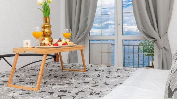 A room with a bed, a wooden table with breakfast items, a vase with flowers, and a view of the sea through a window with curtains. - Apartments Tino (Sutivan)