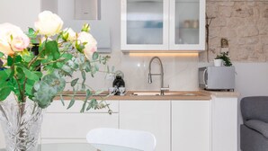 A modern kitchen with white cabinets, a marble countertop, a stainless steel sink, and a microwave. There is a vase with flowers on the counter and a gray sofa in the background. - Apartments Tino (Sutivan)