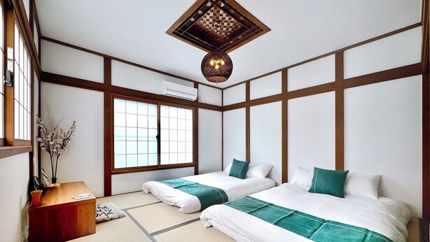 Une pièce de style japonais traditionnel, avec un sol recouvert de tatamis, deux lits dotés de draps verts et blancs, un plafond en bois agrémenté d’un luminaire suspendu et une fenêtre offrant une vue sur la mer.