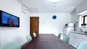 Une salle de conférence avec une longue table, quatre chaises, une horloge, un téléviseur fixé au mur et un coin cuisine.