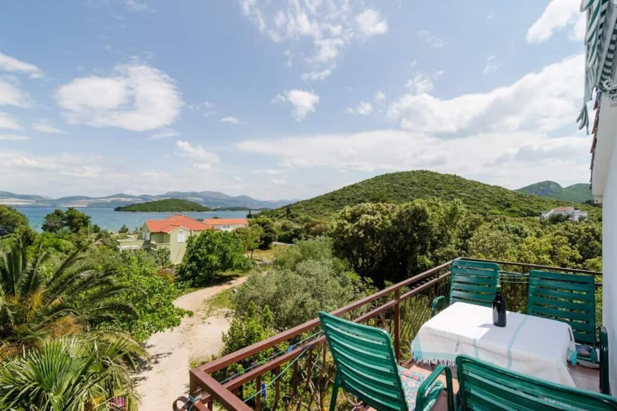 A balcony with a table and chairs overlooking a beach and a mountainous landscape.