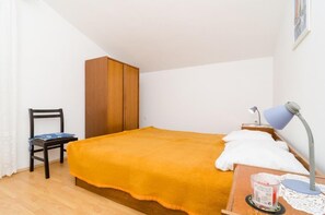 A bedroom with a bed, a wooden wardrobe, a chair, a desk with a lamp, and a small table with a cup.