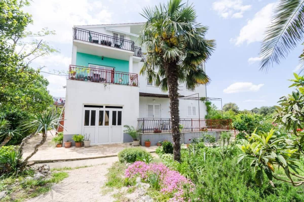 A two-story building with a balcony on the upper floor, surrounded by lush greenery and flowering plants.