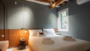 A bedroom with a wooden headboard, a bed with white linens, a bedside table with a lamp, and a window with a view. - Venice Jewel Canal View by Wonderful Italy (Venice)