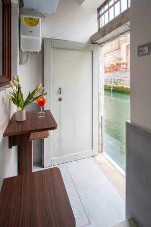 A room with a wooden table, a potted plant, and a glass with a drink. A white door leads to a view of a water body. - Venice Jewel Canal View by Wonderful Italy (Venice)