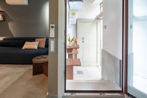 A modern living room with a sofa, a coffee table, and a small wooden side table. A door leads to a hallway with a small plant and a wall-mounted light fixture. - Venice Jewel Canal View by Wonderful Italy (Venice)