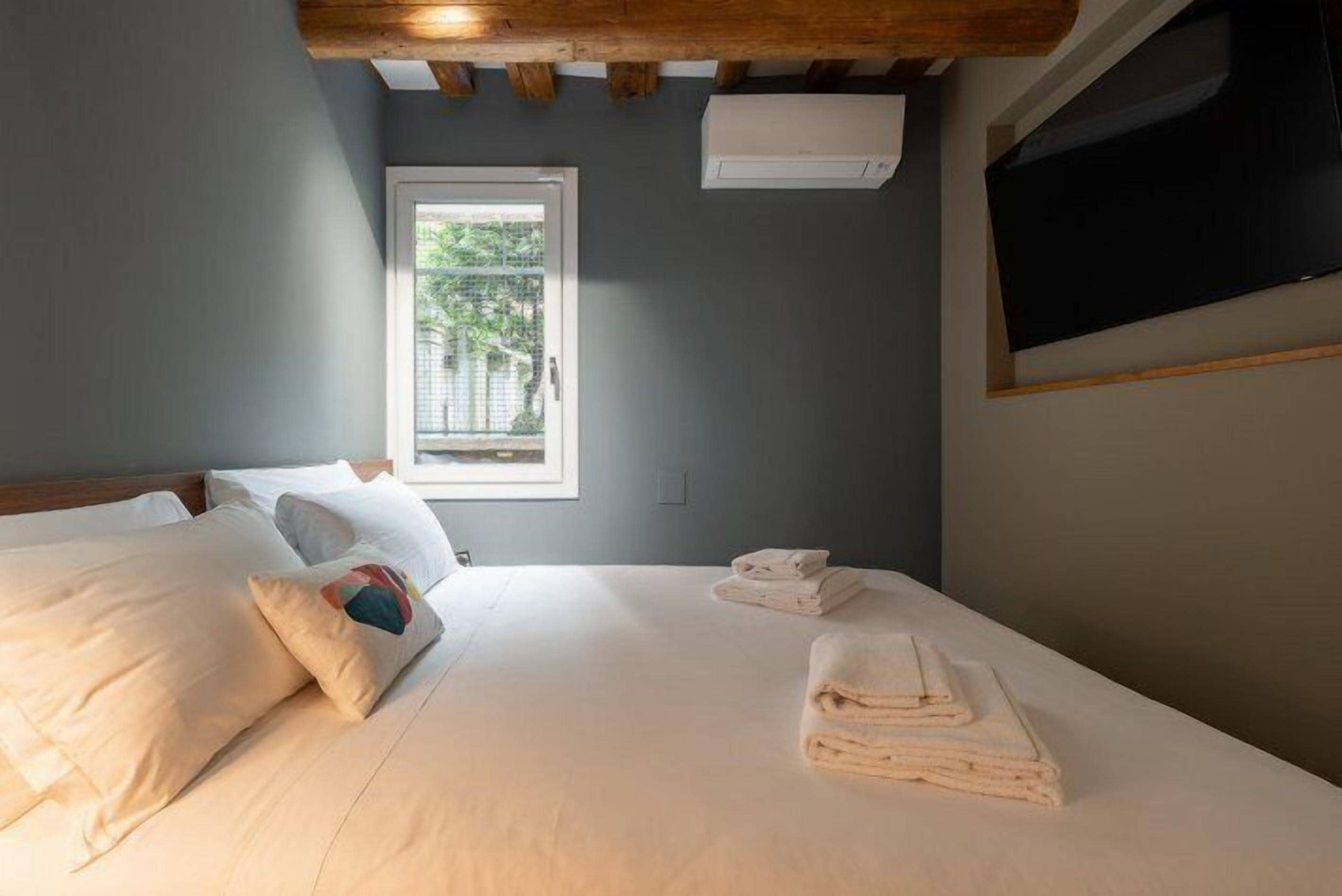 Une chambre à coucher avec un lit, des oreillers, une fenêtre, un climatiseur et un téléviseur fixé au mur.
